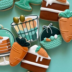 May include: A selection of Easter-themed biscuits. The biscuits feature carrot shapes, cake slices with carrot decorations, and basket-shaped biscuits with eggs. The biscuits are decorated with white, orange, brown, and teal icing.