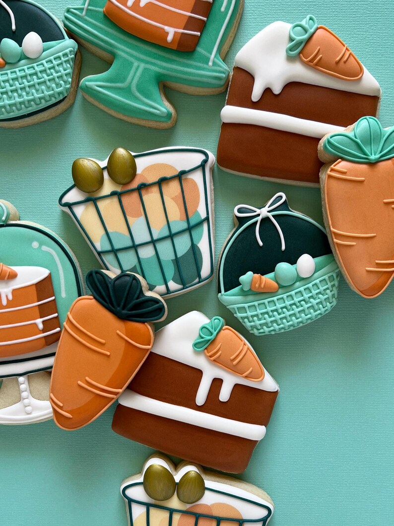 May include: A selection of Easter-themed biscuits. The biscuits feature carrot shapes, cake slices with carrot decorations, and basket-shaped biscuits with eggs. The biscuits are decorated with white, orange, brown, and teal icing.