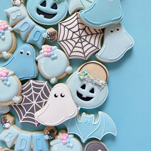 May include: A collection of Halloween-themed sugar cookies decorated with blue, white, and gray icing. The cookies feature ghosts, bats, pumpkins, spiderwebs, and a "BOO" banner.