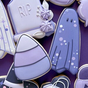 Puede incluir: Una colecci&oacute;n de galletas de az&uacute;car de Halloween de color morado y blanco. Las galletas est&aacute;n decoradas con varios dise&ntilde;os, incluyendo un fantasma, un sombrero de bruja, un ma&iacute;z dulce, una calabaza, una l&aacute;pida con la inscripci&oacute;n "RIP" y una bolsa de regalo.