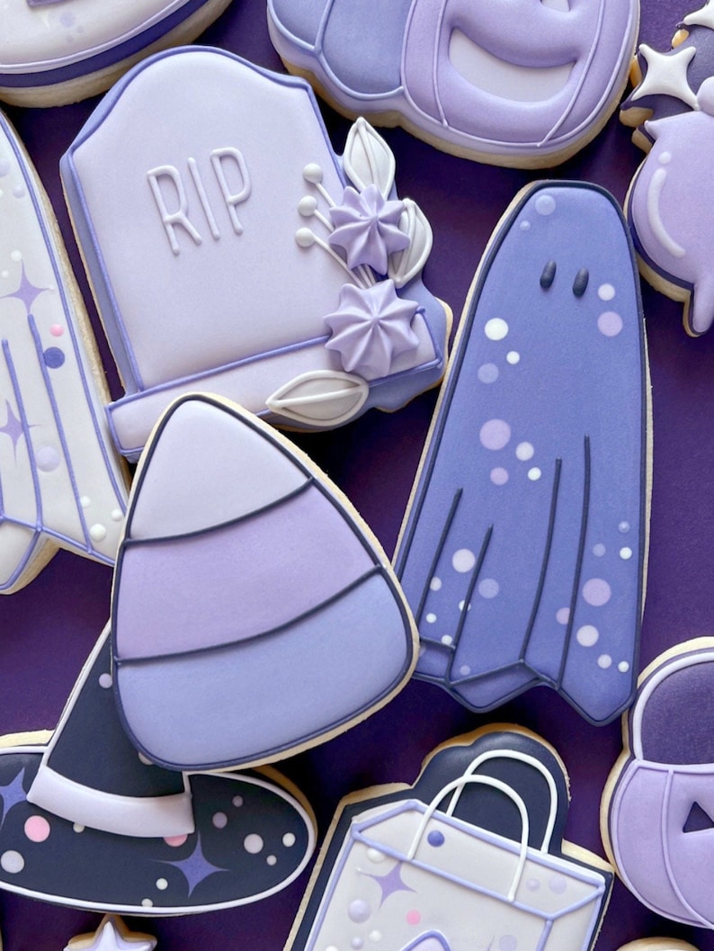 Puede incluir: Una colecci&oacute;n de galletas de az&uacute;car de Halloween de color morado y blanco. Las galletas est&aacute;n decoradas con varios dise&ntilde;os, incluyendo un fantasma, un sombrero de bruja, un ma&iacute;z dulce, una calabaza, una l&aacute;pida con la inscripci&oacute;n "RIP" y una bolsa de regalo.