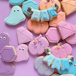May include: A collection of Halloween-themed sugar cookies decorated with various colors and designs. The cookies include ghosts, pumpkins, diamonds, and a banner that reads "BOO".