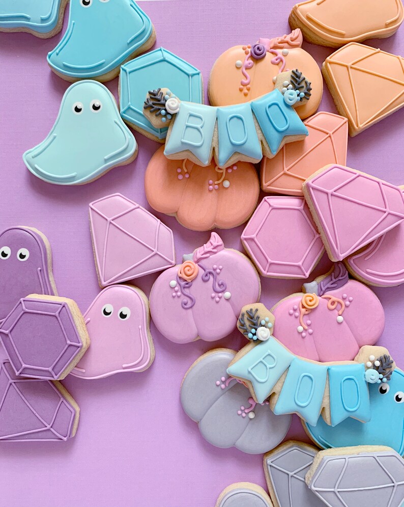 May include: A collection of Halloween-themed sugar cookies decorated with various colors and designs. The cookies include ghosts, pumpkins, diamonds, and a banner that reads "BOO".