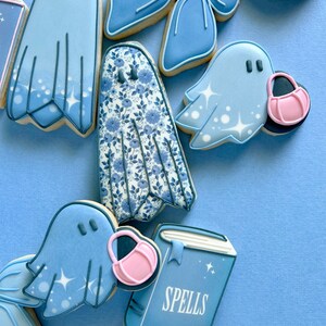 Puede incluir: Una colecci&oacute;n de galletas de az&uacute;car decoradas en azul y blanco con fantasmas, un libro con la palabra "Spells" en la portada, un lazo y un patr&oacute;n floral.