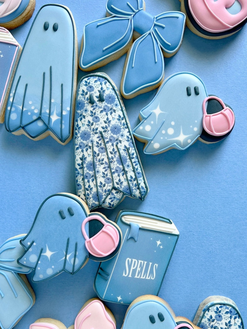 Puede incluir: Una colecci&oacute;n de galletas de az&uacute;car decoradas en azul y blanco con fantasmas, un libro con la palabra "Spells" en la portada, un lazo y un patr&oacute;n floral.
