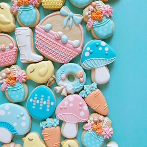 May include: A collection of Easter-themed biscuits on a light blue surface. The biscuits feature designs such as decorated eggs, baskets, mushrooms, carrots, chicks, and a wellington boot. The biscuits are decorated with pastel colours and floral accents.