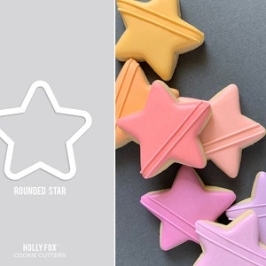 May include: A set of six star-shaped cookie cutters in various shades of pink, orange, and purple. The cutters are made of metal and have a rounded edge. The text "ROUNDED STAR" is printed on a white background.