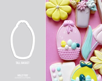 May include: A cookie cutter in the shape of a tall basket with a scalloped top. The cutter is white and is shown on a grey background. The text "Tall Basket" is below the cutter and the text "HOLLY FOX COOKIE CUTTERS" is below that.