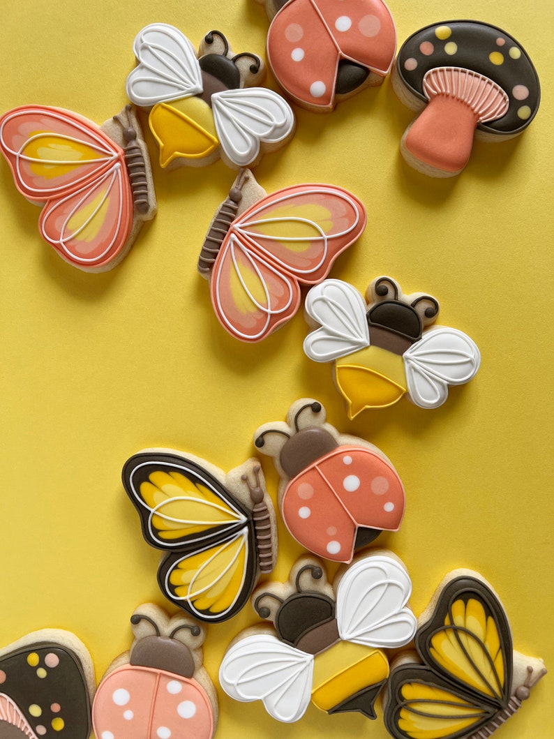 Pode incluir: Uma cole&ccedil;&atilde;o de biscoitos de a&ccedil;&uacute;car coloridos decorados com v&aacute;rios desenhos tem&aacute;ticos da natureza, incluindo borboletas, joaninhas, abelhas e cogumelos. Os biscoitos est&atilde;o dispostos em um fundo amarelo.