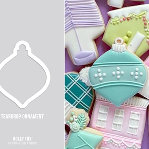 May include: A white teardrop-shaped cookie cutter for making Christmas ornaments. The cutter is shown against a gray background. The text "Teardrop Ornament" and "Holly Fox Cookie Cutters" is printed below the cutter.