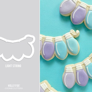 May include: A cookie cutter in the shape of a string of Christmas lights. The cutter is made of metal and has a white finish. The cutter is shown on a gray background. The text "Light String" is printed below the cutter.