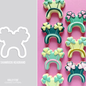 May include: A collection of shamrock headband cookies with green, pink, and yellow icing. The cookies are decorated with shamrock designs and are arranged on a pink background. The image also shows a white cookie cutter with the text "SHAMROCK HEADBAND".