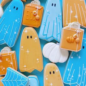 Puede incluir: Una colecci&oacute;n de galletas de az&uacute;car con tem&aacute;tica de Halloween decoradas con glaseado azul y naranja. Las galletas tienen forma de fantasmas, calabazas y bolsas de la compra. Algunas de las galletas tienen purpurina. Las galletas est&aacute;n dispuestas sobre un fondo azul.