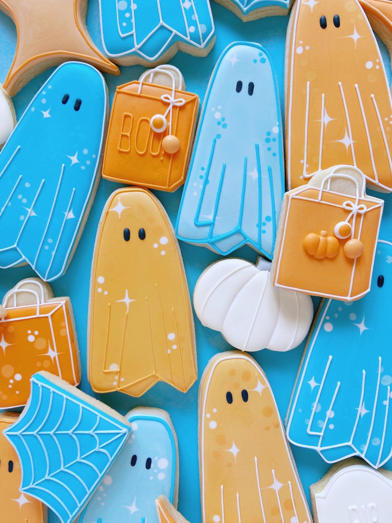 Puede incluir: Una colecci&oacute;n de galletas de az&uacute;car con tem&aacute;tica de Halloween decoradas con glaseado azul y naranja. Las galletas tienen forma de fantasmas, calabazas y bolsas de la compra. Algunas de las galletas tienen purpurina. Las galletas est&aacute;n dispuestas sobre un fondo azul.