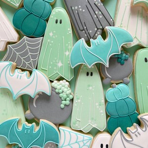 Puede incluir: Una colecci&oacute;n de galletas de az&uacute;car con tem&aacute;tica de Halloween decoradas con glaseado verde, blanco y gris. Las galletas tienen forma de fantasmas, murci&eacute;lagos, calabazas y calderos.