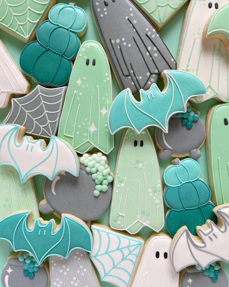 Puede incluir: Una colecci&oacute;n de galletas de az&uacute;car con tem&aacute;tica de Halloween decoradas con glaseado verde, blanco y gris. Las galletas tienen forma de fantasmas, murci&eacute;lagos, calabazas y calderos.