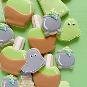 May include: A collection of Halloween-themed sugar cookies decorated with green, brown, and gray icing. The cookies are shaped like ghosts, cauldrons, potion bottles, and candy apples.