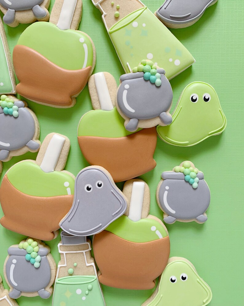 May include: A collection of Halloween-themed sugar cookies decorated with green, brown, and gray icing. The cookies are shaped like ghosts, cauldrons, potion bottles, and candy apples.