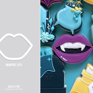 May include: A white cookie cutter in the shape of lips, with the text "VAMPIRE LIPS" below it. The image also shows decorated cookies, including a purple vampire mouth with fangs, and other Halloween-themed shapes in blue and yellow.