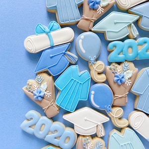 May include: A collection of blue and white graduation themed sugar cookies. The cookies are decorated with images of graduation caps, diplomas, gowns, and balloons. The cookies also feature the year 2020.