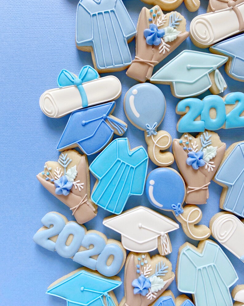 May include: A collection of blue and white graduation themed sugar cookies. The cookies are decorated with images of graduation caps, diplomas, gowns, and balloons. The cookies also feature the year 2020.