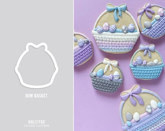 May include: A set of six cookie cutters shaped like Easter baskets with bows. The cutters are made of white plastic and are shown on a purple background. The text "BOW BASKET" is printed below the cutters.
