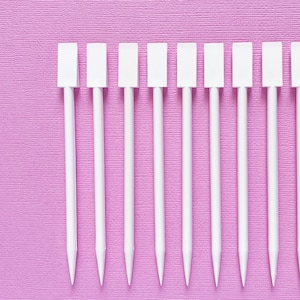 Peut inclure: Vue de dessus d'une rangée de brochettes blanches à dessus rectangulaires sur un fond texturé, violet clair. Les brochettes sont espacées uniformément et ont une extrémité pointue. Le fond est de couleur violet clair uni.