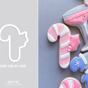 Pode incluir: Um cortador de biscoitos branco em forma de bengala de caramelo com um ramo de azevinho. O cortador est&aacute; sobre um fundo cinza. O texto "CANDY CANE W/ SPRIG" est&aacute; abaixo do cortador. O texto "HOLLY FOX" e "COOKIE CUTTERS" est&aacute; abaixo do texto.