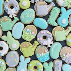 May include: A collection of Easter-themed sugar cookies decorated with pastel colors and various designs, including bunnies, eggs, watering cans, baskets, and jelly beans.