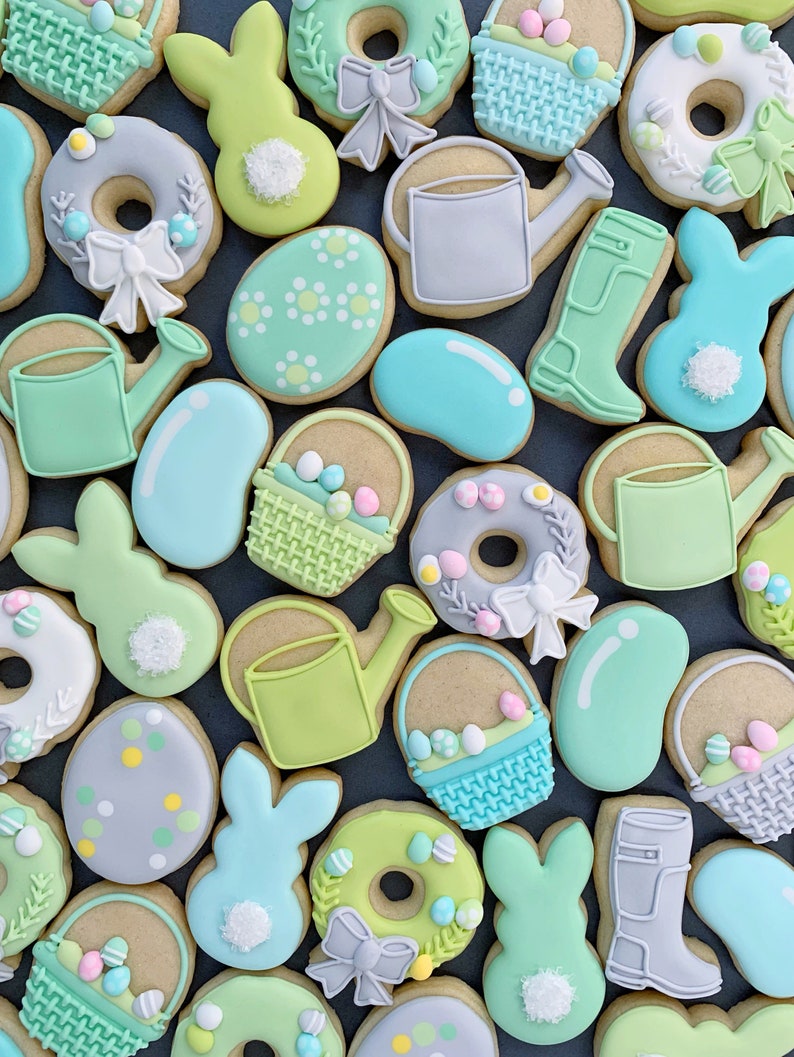 May include: A collection of Easter-themed sugar cookies decorated with pastel colors and various designs, including bunnies, eggs, watering cans, baskets, and jelly beans.