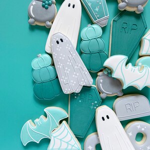 Puede incluir: Una colecci&oacute;n de galletas de az&uacute;car con tem&aacute;tica de Halloween decoradas con glaseado de color verde azulado, blanco y gris. Las galletas representan fantasmas, murci&eacute;lagos, calabazas, calderos y l&aacute;pidas con la inscripci&oacute;n "RIP".