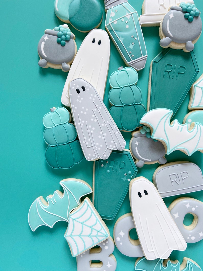 Puede incluir: Una colecci&oacute;n de galletas de az&uacute;car con tem&aacute;tica de Halloween decoradas con glaseado de color verde azulado, blanco y gris. Las galletas representan fantasmas, murci&eacute;lagos, calabazas, calderos y l&aacute;pidas con la inscripci&oacute;n "RIP".