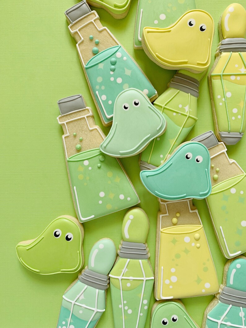 May include: A collection of decorated sugar cookies in the shape of ghosts and potion bottles. The cookies are decorated with green, yellow, and white icing and have a whimsical, spooky theme.