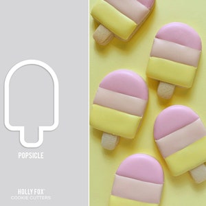 May include: A white popsicle-shaped cookie cutter next to several decorated cookies. The cookies are shaped like popsicles with pink, light pink, and yellow layers, and a wooden stick. The text "POPSICLE" and "HOLLY FOX COOKIE CUTTERS" are visible.