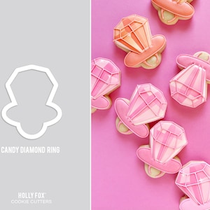 May include: A set of pink and orange cookie cutters shaped like diamond rings. The cookie cutters are on a pink background. The text "CANDY DIAMOND RING" is printed below the cookie cutters. The text "HOLLY FOX COOKIE CUTTERS" is printed below the text "CANDY DIAMOND RING".