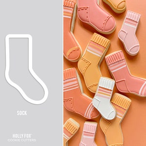 May include: A set of cookie cutters shaped like socks in various colors, including pink, orange, and yellow. The cookie cutters are arranged on a peach-colored background. The text "HOLLY FOX" and "COOKIE CUTTERS" is printed below the cookie cutters.