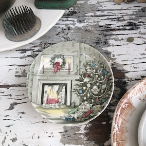 May include: A small, round decorative plate with a Christmas scene featuring a fireplace, Christmas tree, and wrapped gifts. The plate has a gray and white color scheme with a detailed illustration. Other plates and flower frogs are also visible.