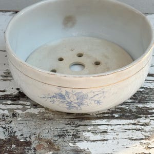 May include: A round, off-white ceramic bowl featuring a delicate blue floral transfer pattern on the exterior and a thin gold rim. The interior contains a removable circular ceramic insert with five drainage holes, showing signs of age.