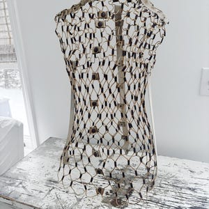May include: A decorative wire mesh form in the shape of a torso. The form is constructed with a beige wire netting, black accents, and small, dark-colored fasteners. It stands on a distressed white surface, suggesting a vintage or antique aesthetic.