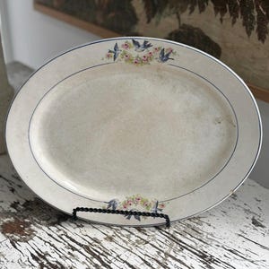 May include: Oval serving platter with a cream-colored surface, featuring a blue border and delicate floral and bird motifs. The vintage-style platter is supported by a black metal stand, perfect for displaying food or as a decorative piece.