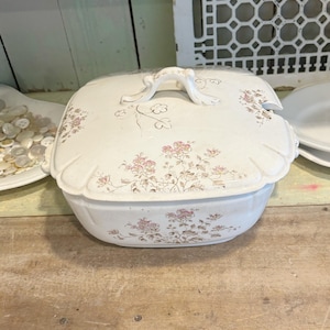 May include: A white ceramic serving dish with a lid, adorned with pink floral patterns. The dish has a curved shape and a decorative handle on the lid. The design is vintage and elegant, suitable for serving food.