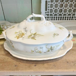 May include: A white porcelain tureen with a lid and matching base. The tureen features a floral design in shades of yellow and green. The lid has a branch-shaped handle. The tureen is sitting on a wooden surface.