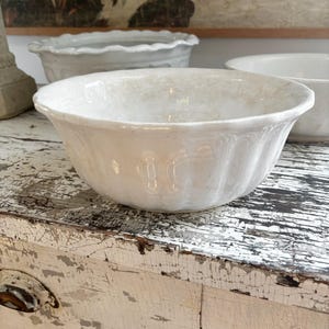 May include: A white ceramic bowl with a fluted design. The bowl has a wide rim and sits on a distressed white wooden surface. Other white ceramic bowls are visible in the background, along with a painting.