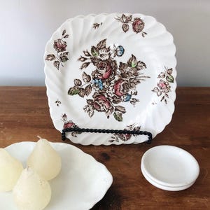 May include: White square plate with a floral rose design in shades of pink, brown, and blue. The plate has a scalloped edge and is displayed on a black metal stand. Additional white plates and pear-shaped candles are also visible.