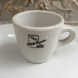 May include: A white ceramic mug with a small handle and a black graphic print on the front. The print features a square outline containing the text GOOD THINGS TO EAT, the words White Tower, and a small illustration of a building with a tower.