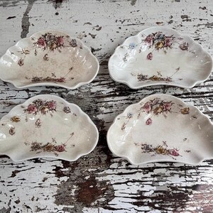 May include: A set of four off-white ceramic dishes with scalloped edges and a floral pattern featuring pink, yellow, and blue roses. Each dish has a glossy finish with a subtle crackle texture and a curved, shell-like shape.