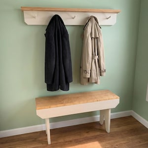 May include: A wooden bench with a light-colored top and white base sits below a wall-mounted coat rack. Two coats hang from the rack: a dark gray coat and a tan trench coat. The wall is painted a light green.