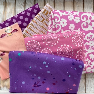 May include: A collection of folded fabric pieces in various patterns and colors. The fabrics include a light peach with dragonfly designs, a deep purple with star and bird motifs, and pink fabrics with floral patterns. The fabrics are arranged on a white wooden surface.