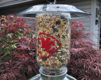 Könnte beinhalten: Ein Vogelhäuschen aus klarem Glas mit einem Metallsockel und einem durchsichtigen Plastikdeckel. Der Futterspender ist mit Vogelfutter gefüllt und hat ein rotes und weißes "KC"-Logo an der Seite.