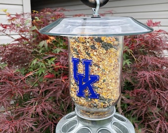 May include: A clear glass bird feeder with a metal base and a blue and white "UK" logo. The feeder is filled with birdseed and is hanging from a hook.